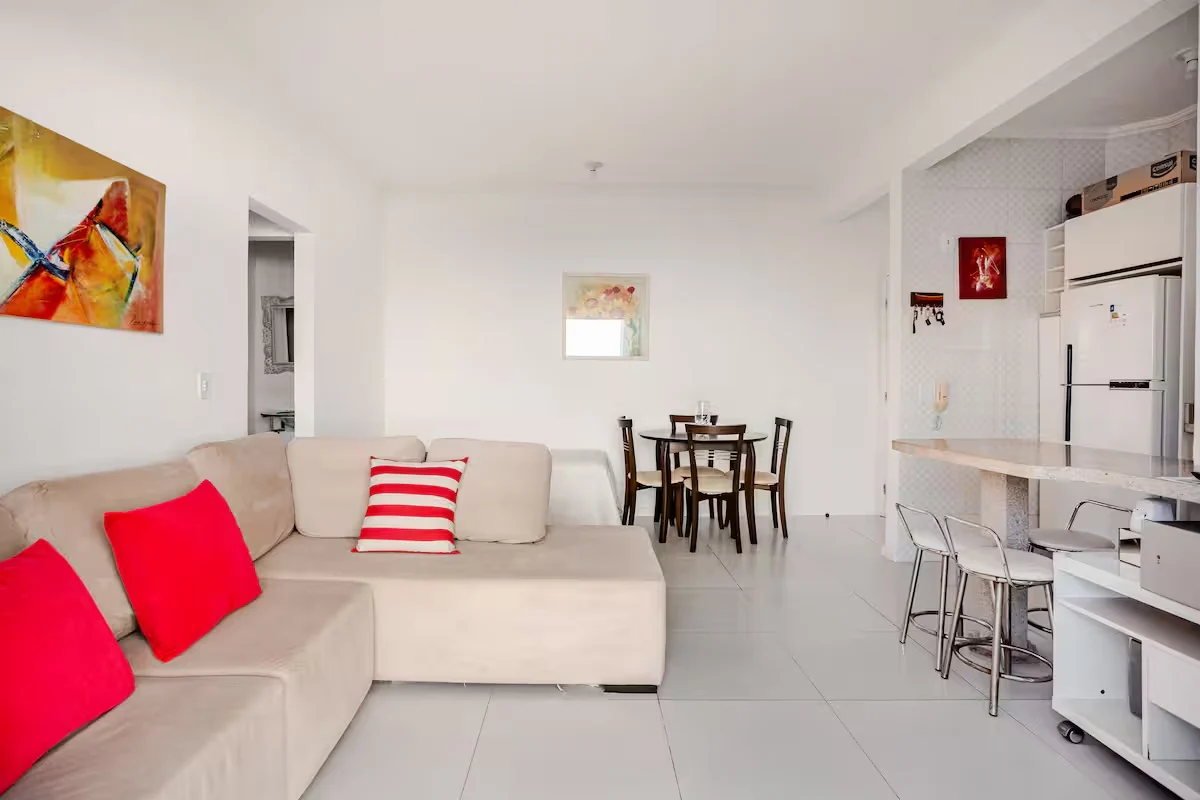 Sala clara com sofá em L, almofadas vermelhas, mesa de jantar e cozinha integrada